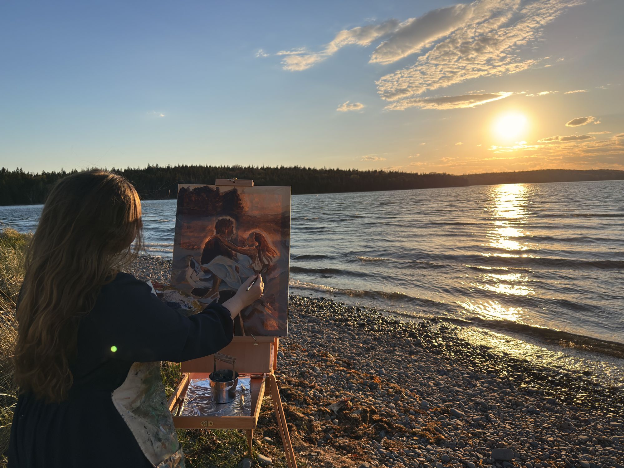 Heather LeBlanc at her easel on a Nova Scotia shoreline at sunset, a finished painting of a couple on the canvas as the sun sets over the water