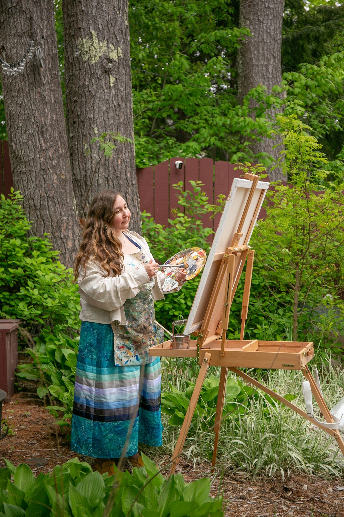 Heather LeBlanc standing at her wooden field easel among the trees, palette in hand, mid-brushstroke