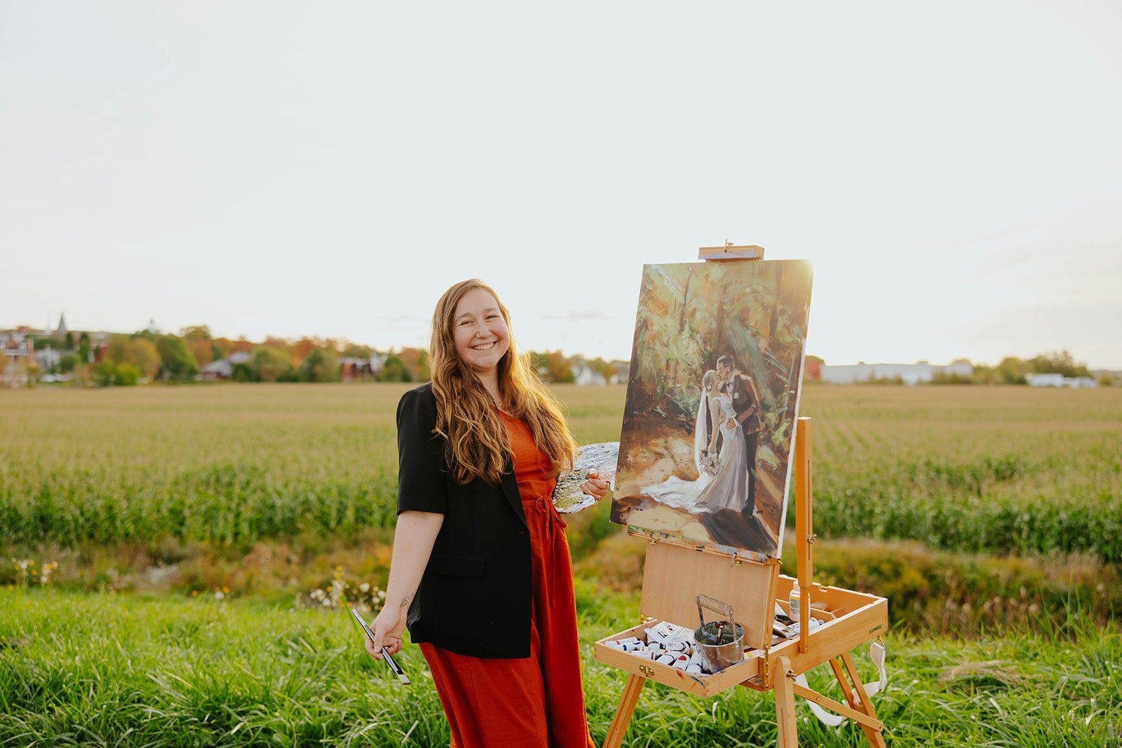 Heather LeBlanc smiling beside her easel in a field at golden hour, palette and brush in hand, with a wedding painting of a couple kissing on the canvas