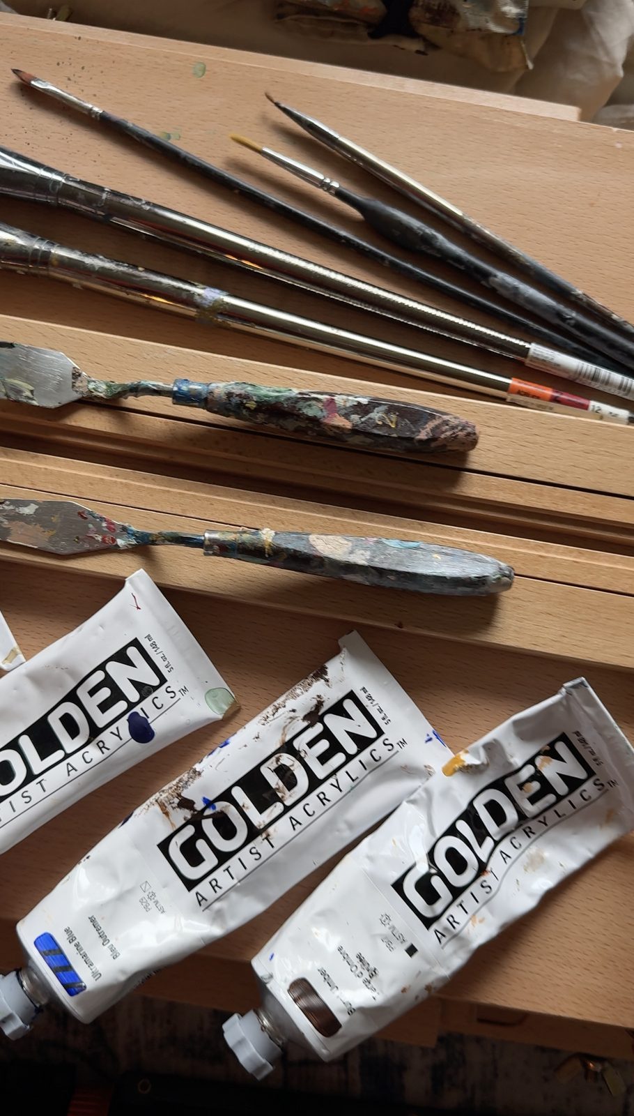 Brushes and tubes of acrylic paint laid out on a wooden table before packing