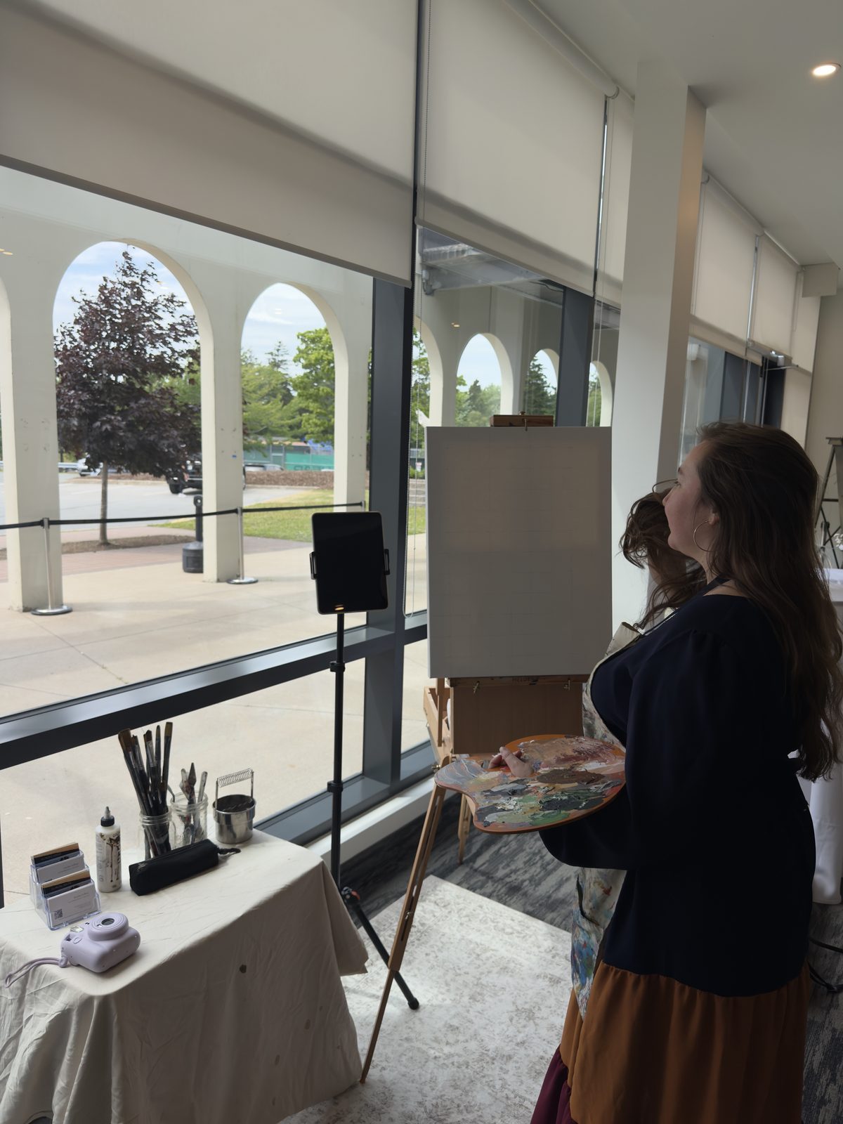 Heather set up with her easel beside a bank of venue windows, palette in hand