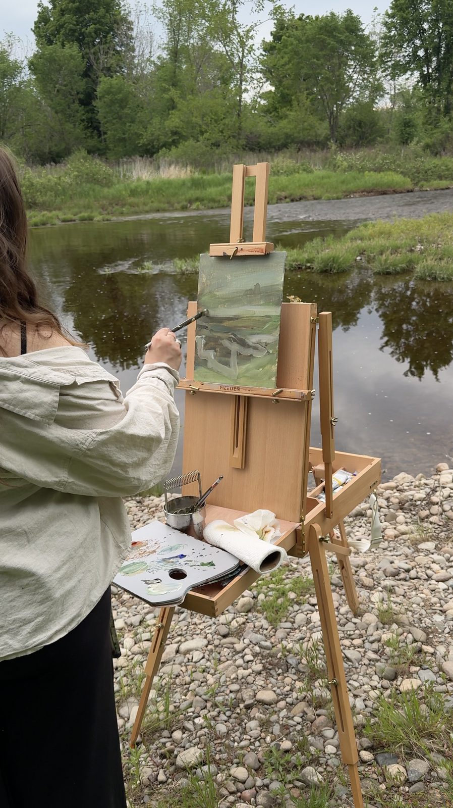 Heather painting a river landscape outdoors, brush in hand, palette on the easel tray