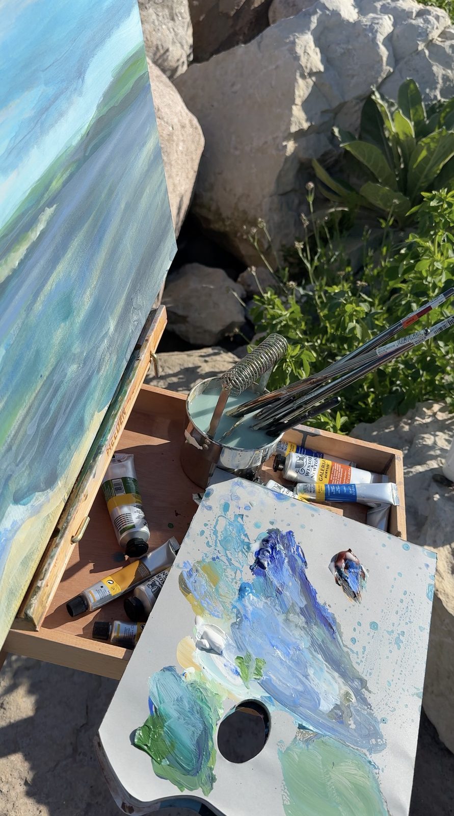 A palette of mixed greens and blues beside the canvas, brushes laid across the paint box
