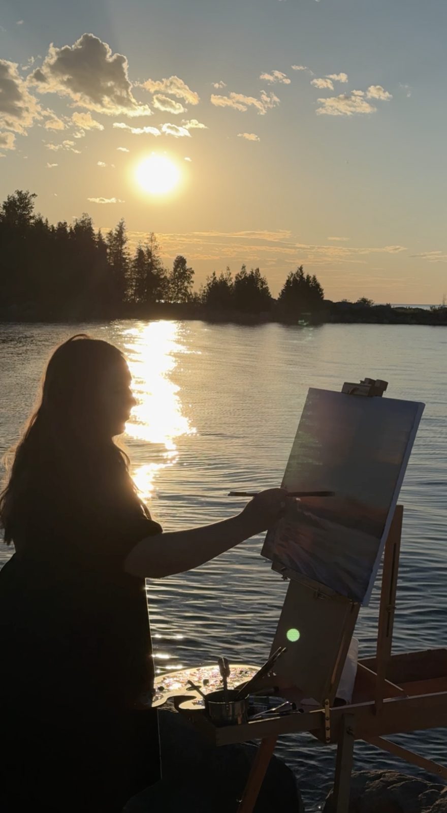 Heather in silhouette at her easel on the shoreline as the sun drops toward the water