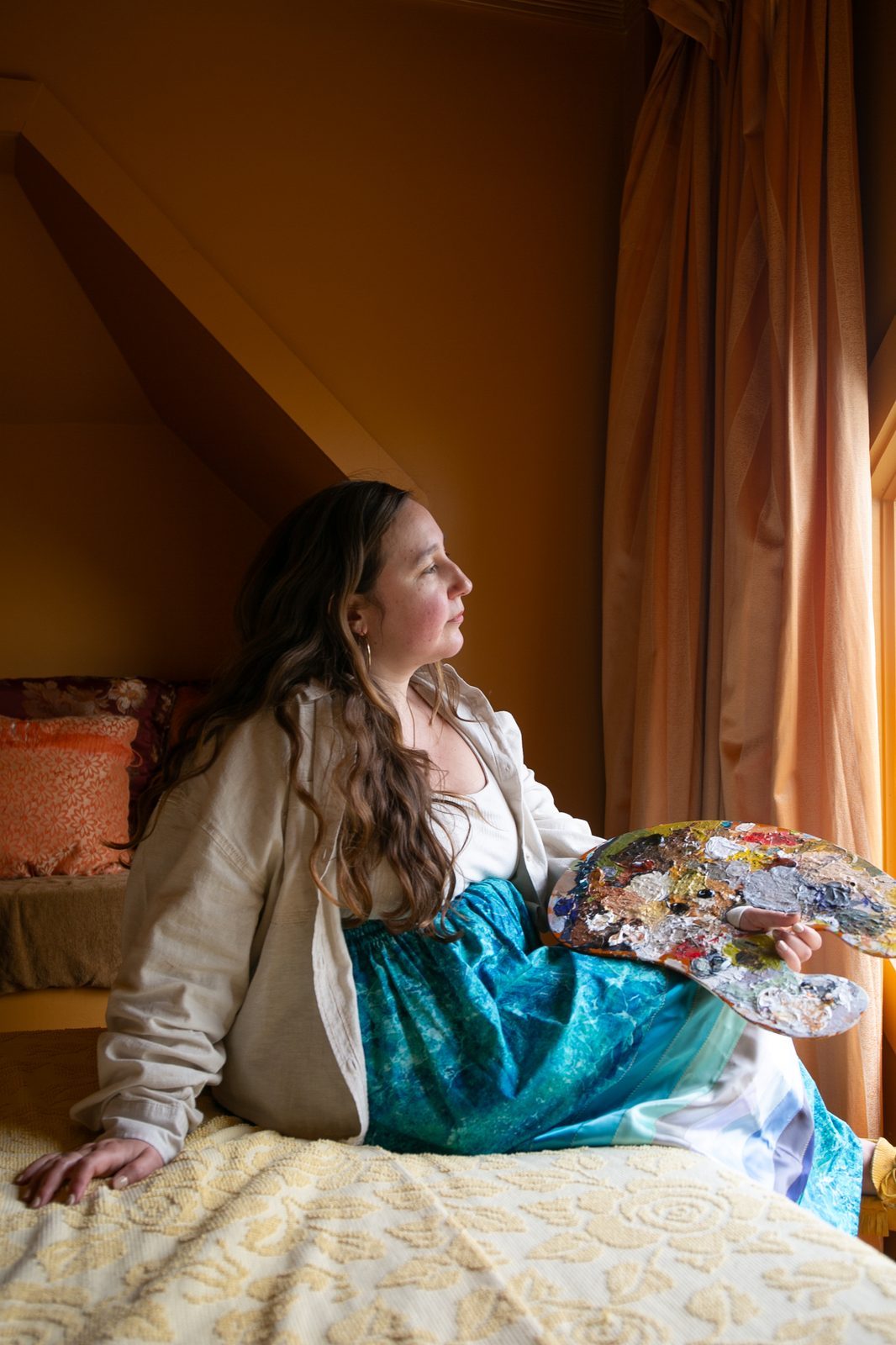 Heather sitting by a window with her palette in her lap, looking out into the light