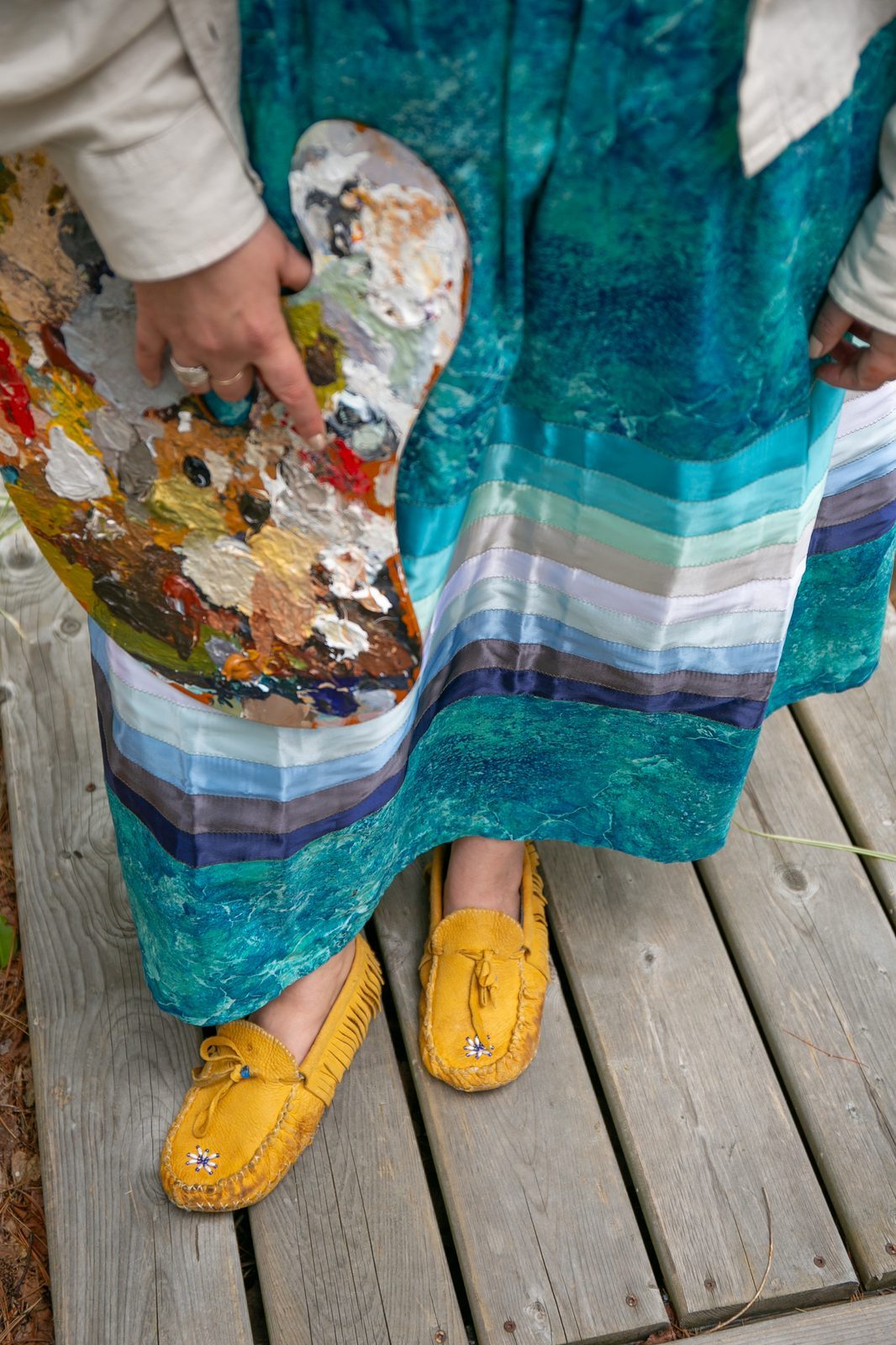 Heather holding her palette, standing on a wooden deck in a ribbon skirt