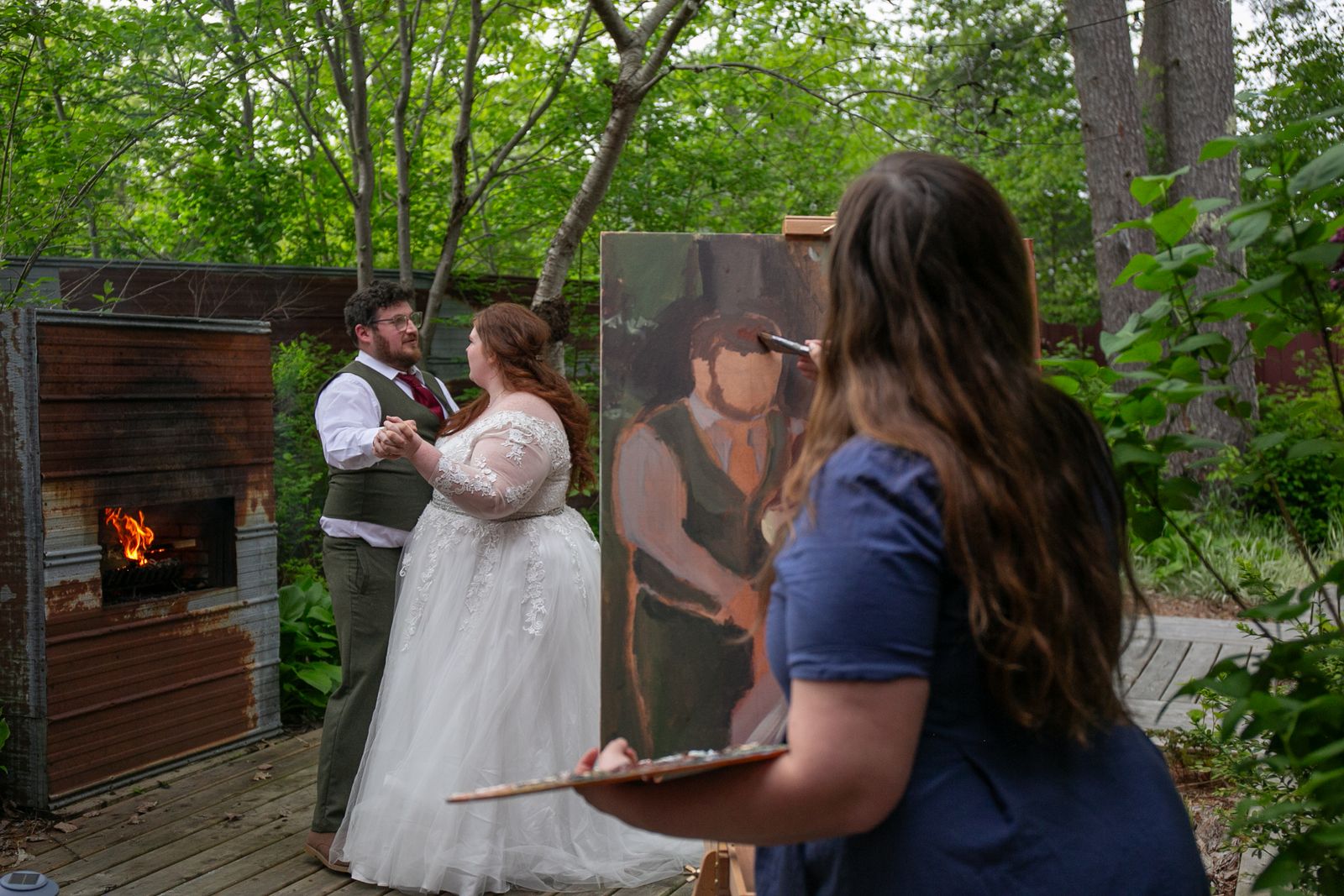 A couple dancing hand in hand beside an outdoor fireplace while Heather paints them live at an easel in the foreground
